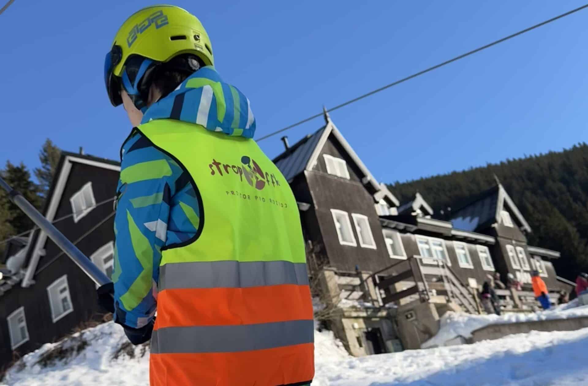 1. Velký obrázek lyžaře s reflexní vestou na sněhu u horských domů.